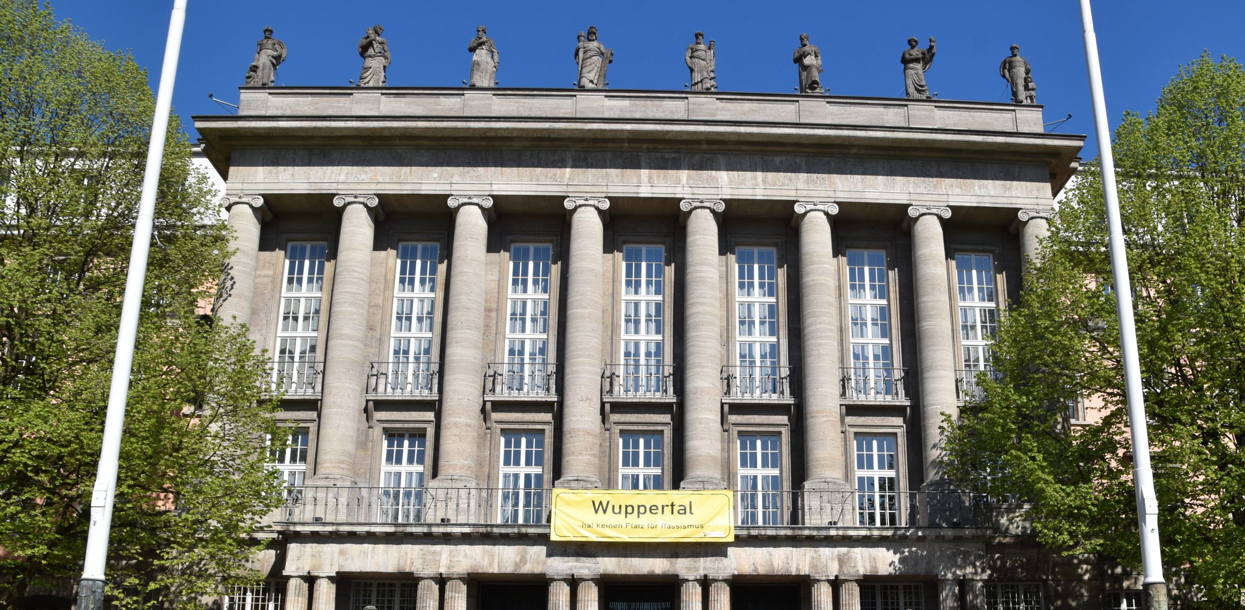 Bild von der Front des Rathauses in Wuppertal Barmen. Daran hängt ein Banner im Stil eines Ortseingangsschildes mit der Aufschrift "Wuppertal hat keinen Platz für Rassismus"